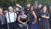 A group of eight people are posing playfully in front of a fence, with each expressing a unique and humorous gesture.