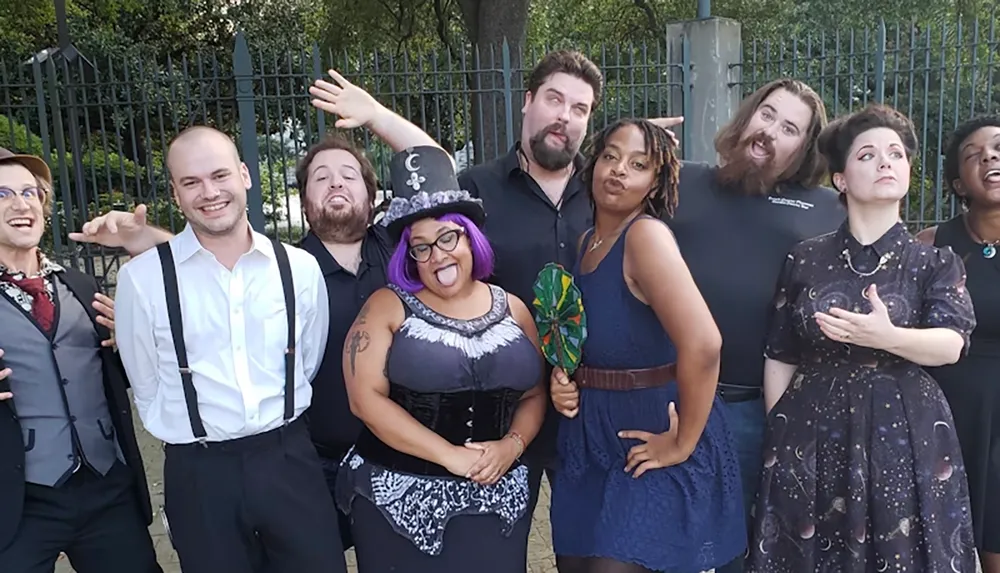 A group of eight people are posing playfully in front of a fence with each expressing a unique and humorous gesture