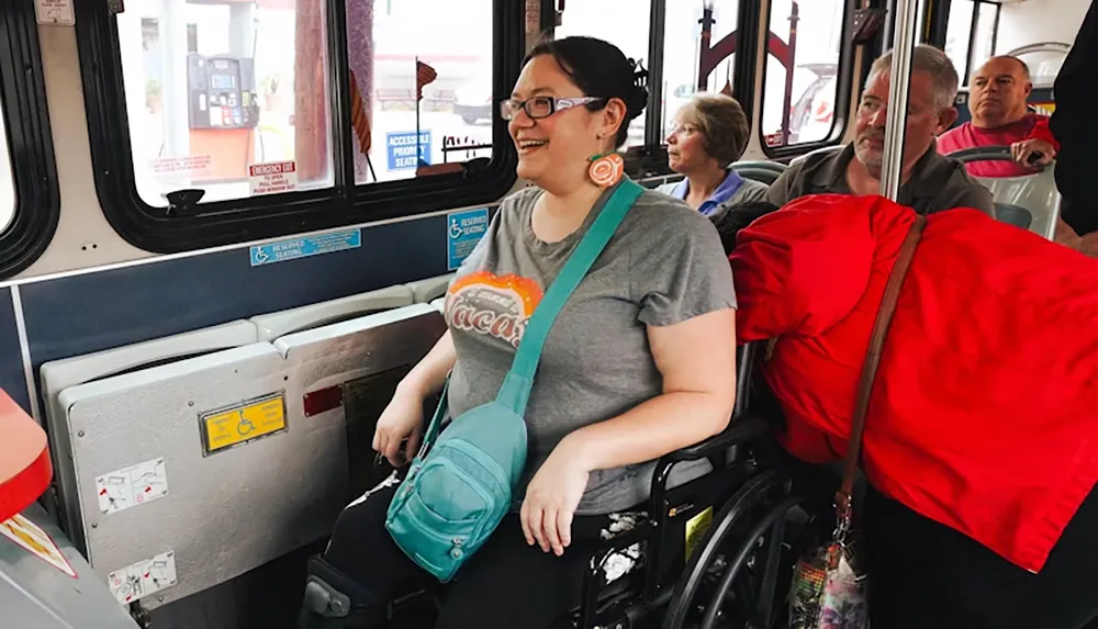 A woman in a wheelchair is smiling while sitting in the designated area on a bus surrounded by other passengers