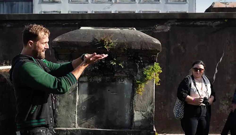 A person stands smiling with outstretched hands next to a stone structure while another person with sunglasses watches nearby