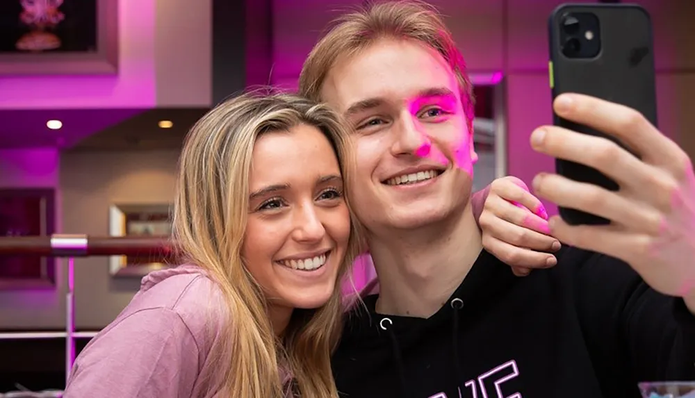 A young man and woman are smiling as they take a selfie together with a pink-lit background