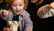 A joyful child eagerly reaches for a dessert with a spoon.