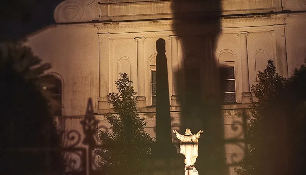 A shadow of a statue with outstretched arms is cast dramatically on the facade of a building at night