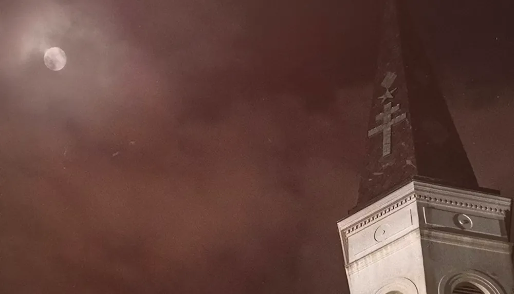 The image shows a church steeple with a cross against a cloudy night sky illuminated by a full moon