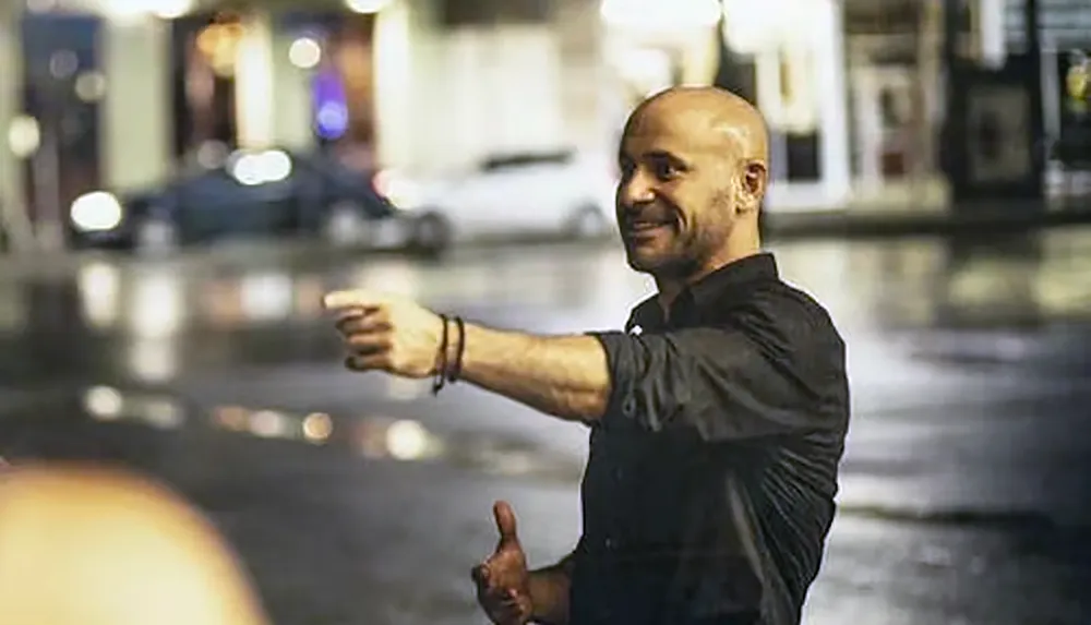 A man is smiling and gesturing with his hand in an urban setting at night