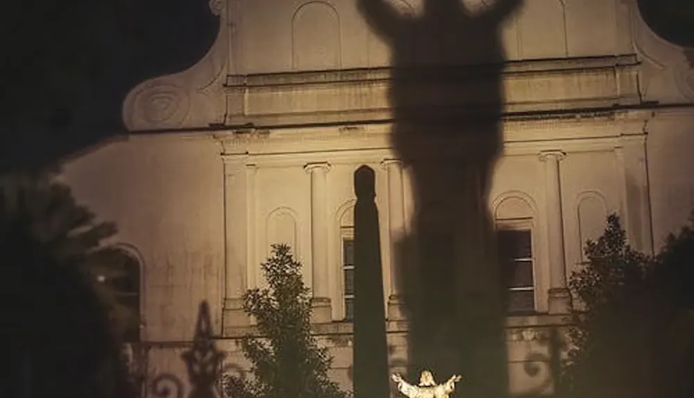 A large shadow of a statue with outstretched arms is projected onto the facade of a building at night