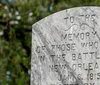 A woman with short white hair wearing a beige shirt gestures while standing in what appears to be a historic cemetery area