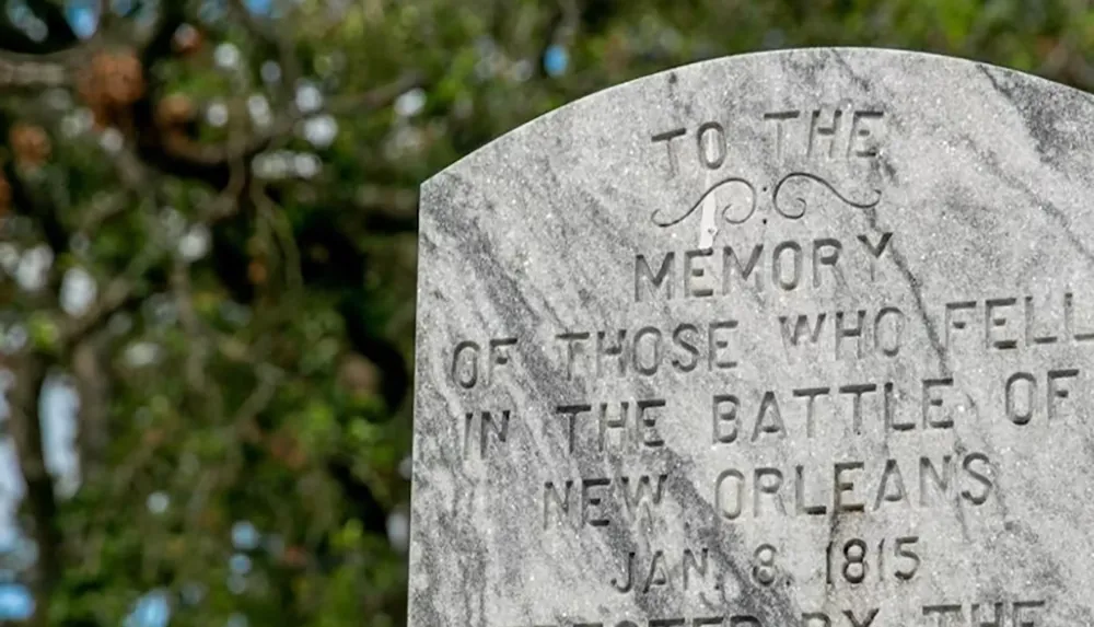 The image shows a weathered tombstone engraved with a dedication to those who fell in the Battle of New Orleans on January 8 1815 set against a blurred background of greenery