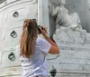 A woman with short white hair wearing a beige shirt gestures while standing in what appears to be a historic cemetery area