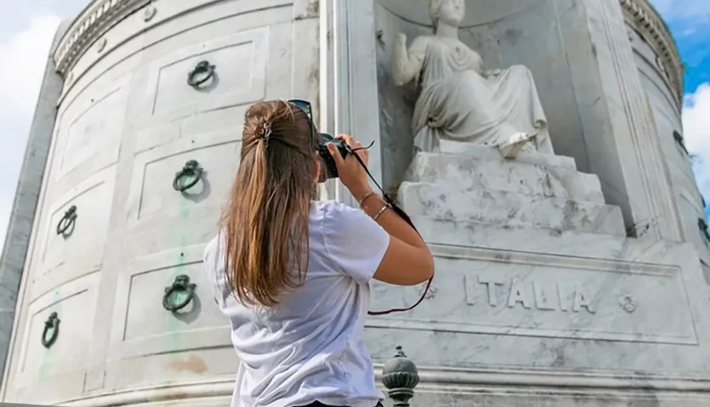 A person in a white shirt is photographing a large stone sculpture marked ITALIA on a monumental building