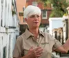 A woman with short white hair wearing a beige shirt gestures while standing in what appears to be a historic cemetery area