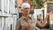 A woman with short white hair, wearing a beige shirt, gestures while standing in what appears to be a historic cemetery area.