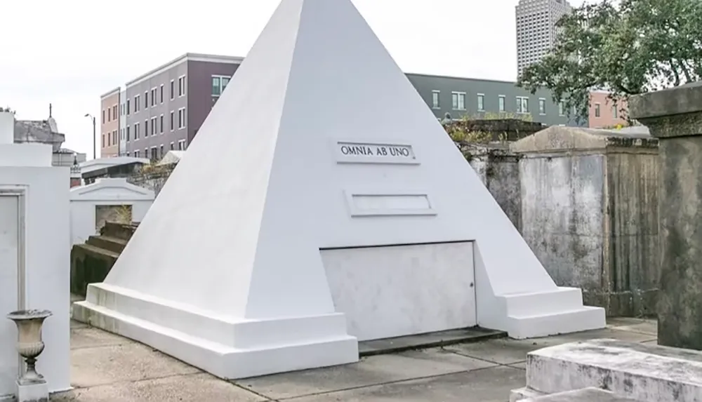 The image shows a white pyramid-shaped tomb with the inscription OMNIA AB UNO in a cemetery setting surrounded by buildings