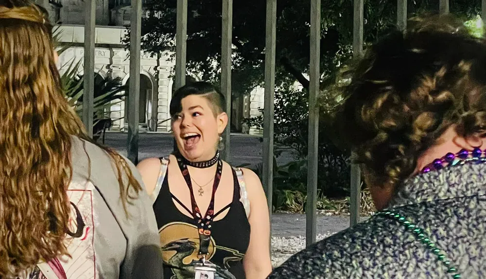 A person with short hair and a choker necklace is smiling and engaging with two other people standing in front of a wrought iron fence at night