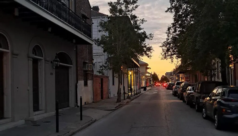 A narrow street lined with parked cars and historic buildings is illuminated by a sunset creating a warm glow in the sky