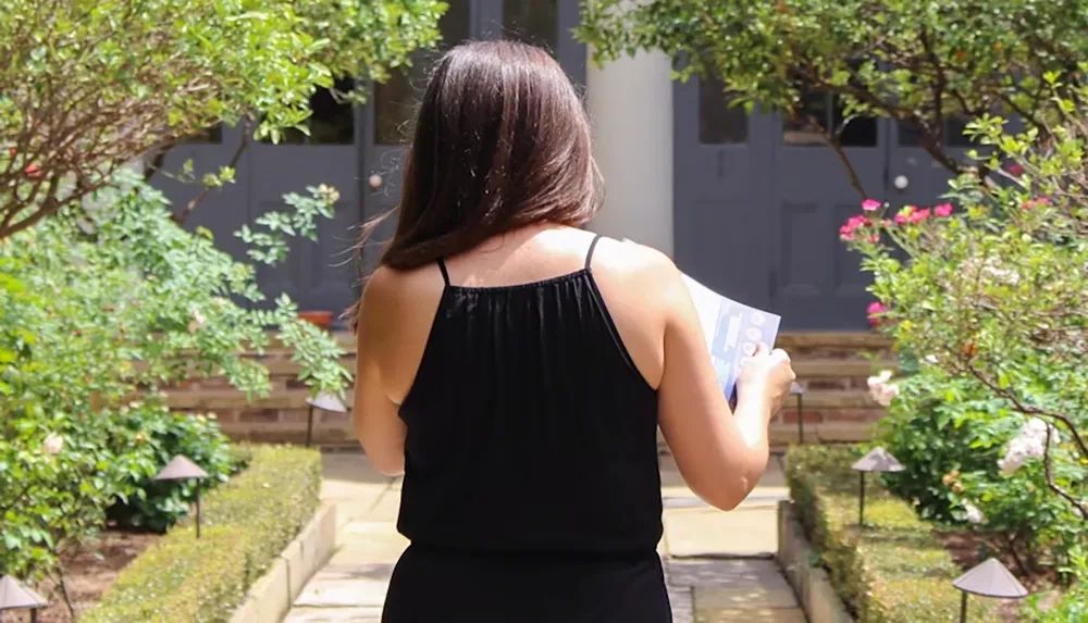 A woman in a black dress is holding a notebook while walking through a garden path towards a house