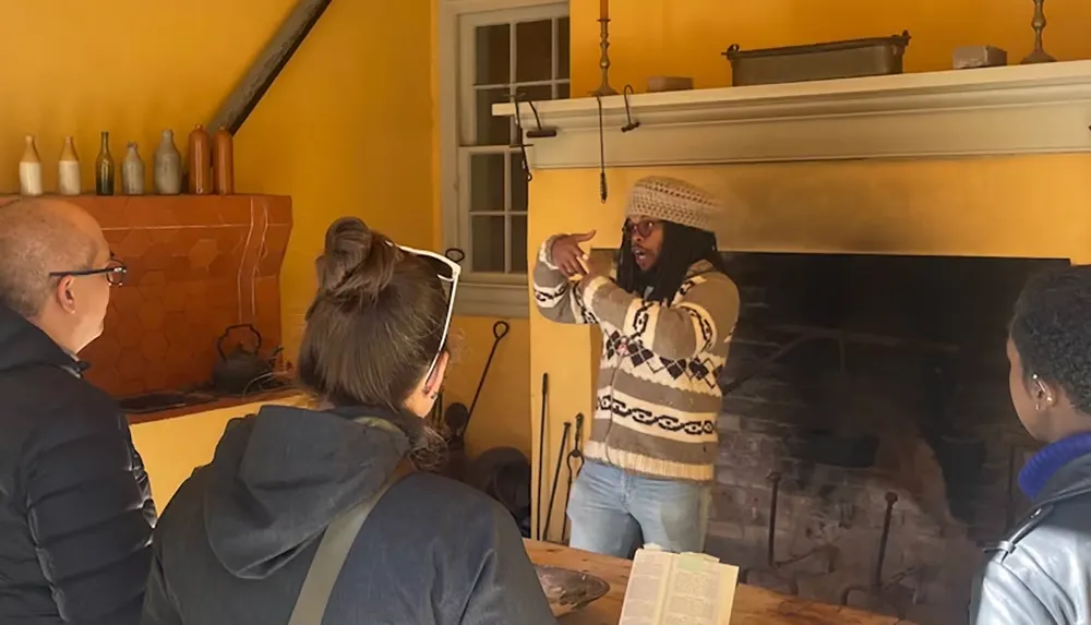 A person in a knit sweater is animatedly gesturing while talking to a group of people inside a cozy rustic room with a large fireplace