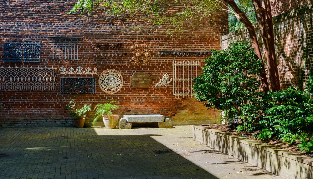 The image shows a sunlit brick courtyard featuring intricate metalwork on the wall potted plants and a stone bench surrounded by lush greenery