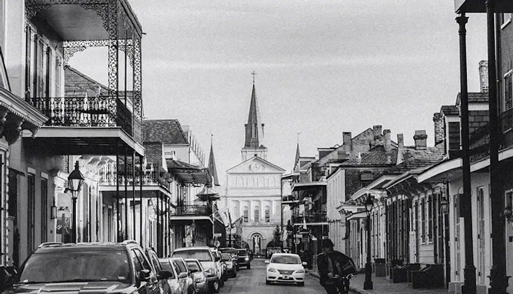 A street scene in black and white showcases a row of historic buildings with ornate balconies lined with parked cars and a church in the background