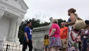 A group of people stands near a white mausoleum, with one person appearing to guide or speak to the others.