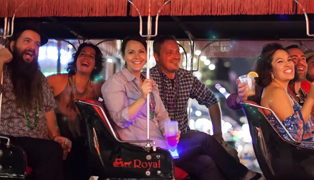 A group of people are joyfully riding a vehicle at night holding drinks and wearing beads