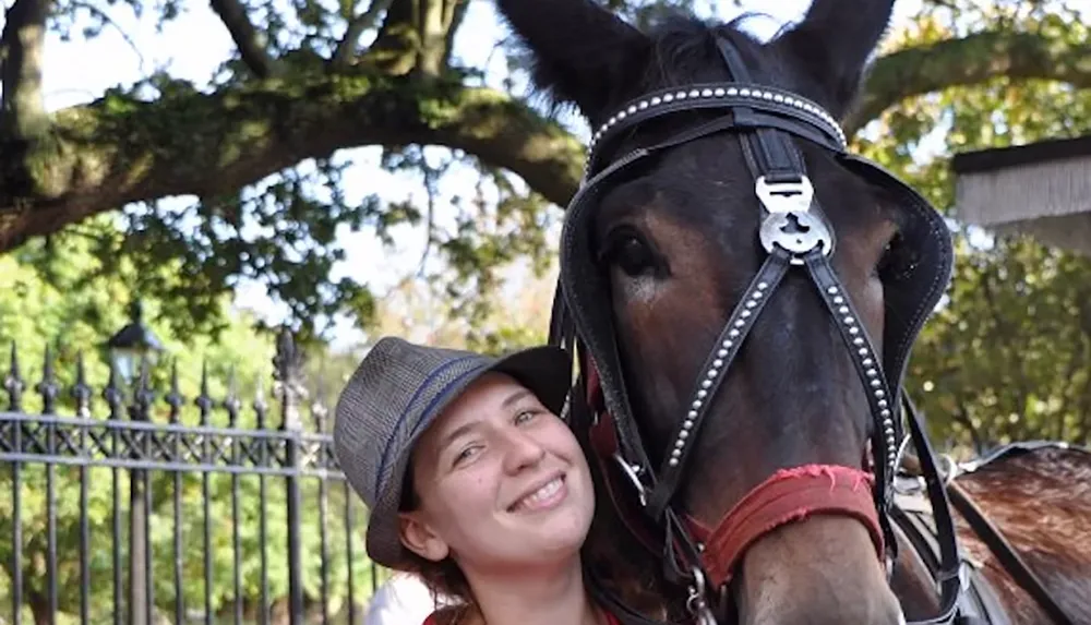 A smiling person wearing a hat stands next to a horse adorned with a harness outdoors