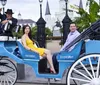 A couple is sitting in a blue horse-drawn carriage with a driver in front of a historic building and lush greenery