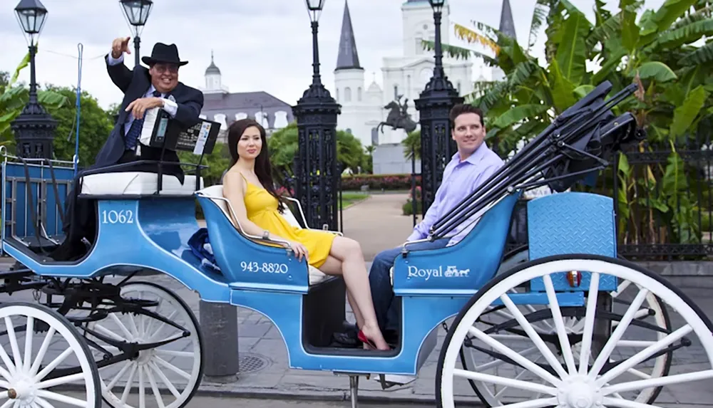 A couple is sitting in a blue horse-drawn carriage with a driver in front of a historic building and lush greenery