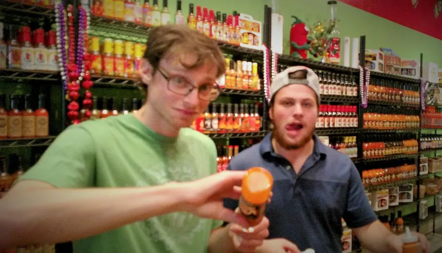 Two people are posing in a store with a wide selection of hot sauces on the shelves in the background.