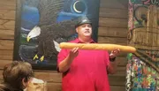 A person in a red shirt is holding a large breadstick in front of an audience, with a painting of an eagle in the background.