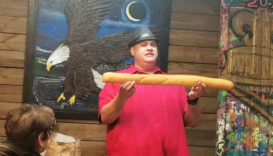 A person in a red shirt is holding a large breadstick in front of an audience, with a painting of an eagle in the background.