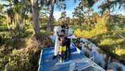 Three people are posing for a photo with smiles on a small blue boat among greenery and water, with tall trees draped with Spanish moss in the background.