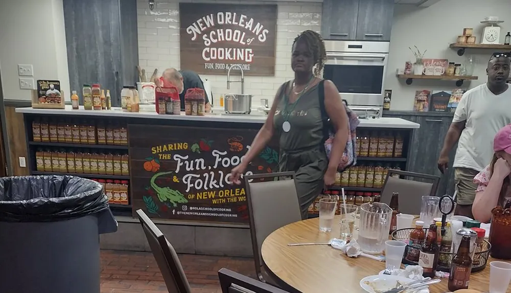 A cooking instructor is standing in front of a class at the New Orleans School of Cooking with participants looking on amidst a setting of dining tables and Southern food products