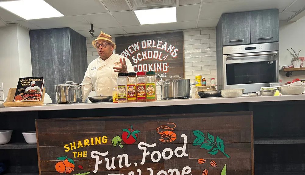 A chef is teaching a cooking class at the New Orleans School of Cooking surrounded by pots spices and a cookbook