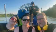 Three people are posing for a selfie in front of a person operating an airboat, with all of them wearing headphones and smiling during a sunny day on the water.