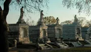 The image shows a series of ornate, stone mausoleums with sculpted figures and crosses in a tranquil cemetery during what appears to be early evening light.