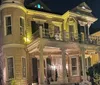 The image shows a two-story corner house at night illuminated by warm lighting featuring classical architectural details such as columns and decorative railings