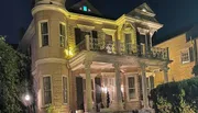 The image shows a two-story corner house at night illuminated by warm lighting, featuring classical architectural details such as columns and decorative railings.