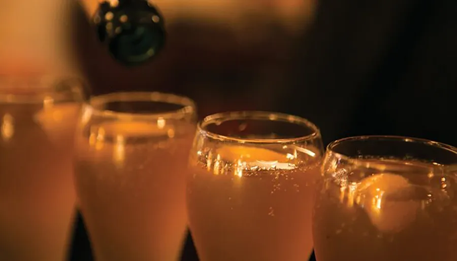 A row of glasses filled with a golden-hued beverage, possibly a cocktail, with a warm ambient lighting setting a cozy atmosphere.