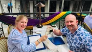 Two people are cheerfully toasting with drinks at an outdoor restaurant adorned with colorful fabrics.