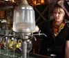 A person wearing a pirate hat is tending a bar with an antique absinthe dispenser filled with chilled water poised to drip over sugar cubes on slotted spoons atop glasses of absinthe