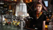 A person wearing a pirate hat is tending a bar with an antique absinthe dispenser filled with chilled water, poised to drip over sugar cubes on slotted spoons atop glasses of absinthe.