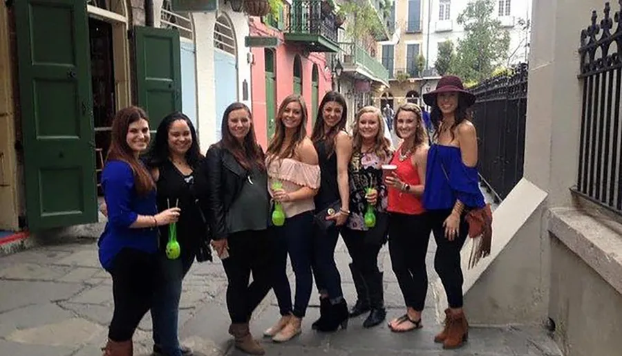 A group of smiling women poses together on a narrow street, some holding beverages, with colorful buildings and a wrought-iron railing in the background.