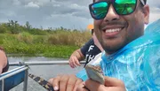 A smiling person is wearing sunglasses and holding a small alligator or crocodile on a boat with water and vegetation in the background.