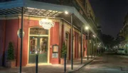 This image captures a tranquil evening scene of an empty street lined with colorful, historic buildings, featuring a restaurant with a lit sign indicating its name and the year it was established.