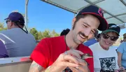 A smiling person in a red shirt and black cap is gently holding a small turtle, with others looking on from a boat with a canopy.