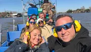 A group of smiling people wearing protective ear muffs are enjoying a ride on an airboat, with waterfront homes visible in the background.