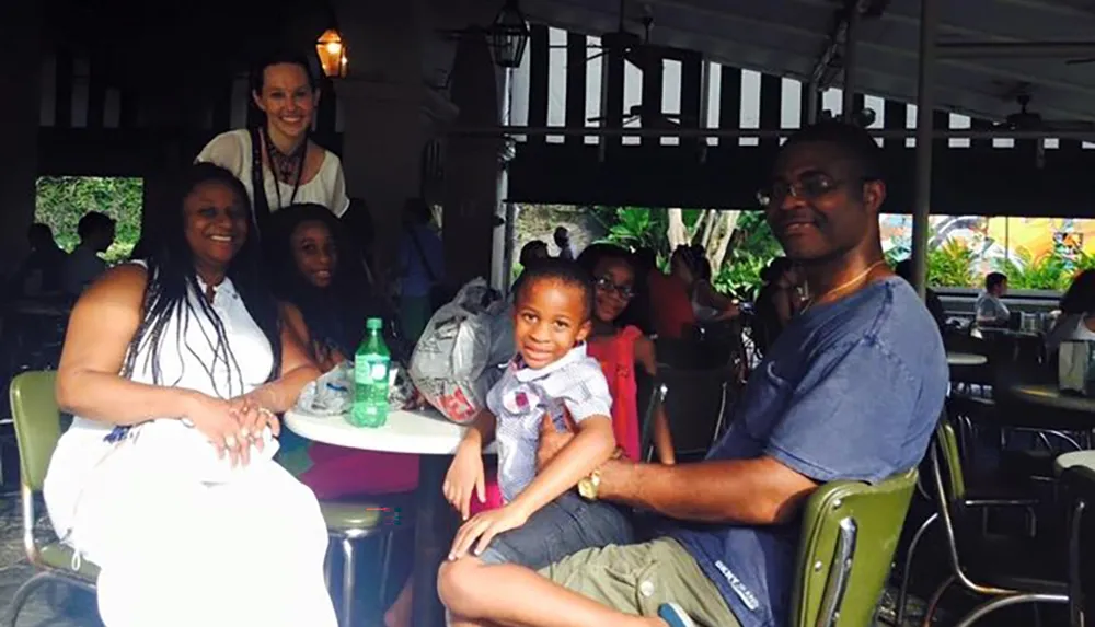 A group of people including a child are smiling and seated at a table in an outdoor dining area with a waitress standing behind them