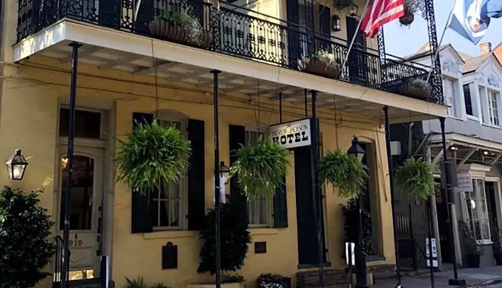 The image shows the facade of a charming yellow building with a balcony adorned with green plants and an American flag identified as a hotel with a sign hanging at the entrance