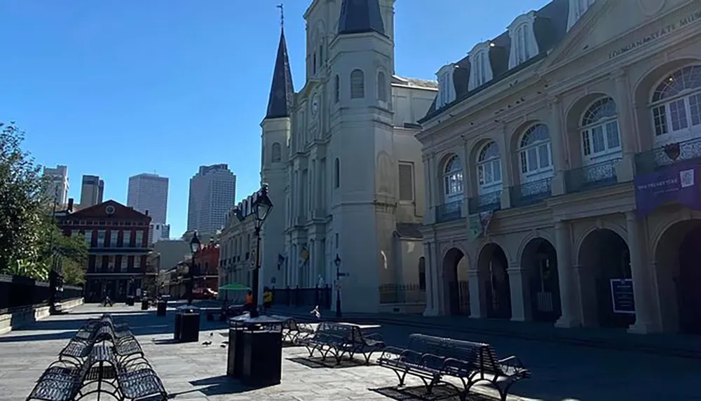 The image shows a sunny empty plaza with benches street lamps and historic architecture under a clear blue sky juxtaposing the old-world charm in the foreground with modern skyscrapers in the distance